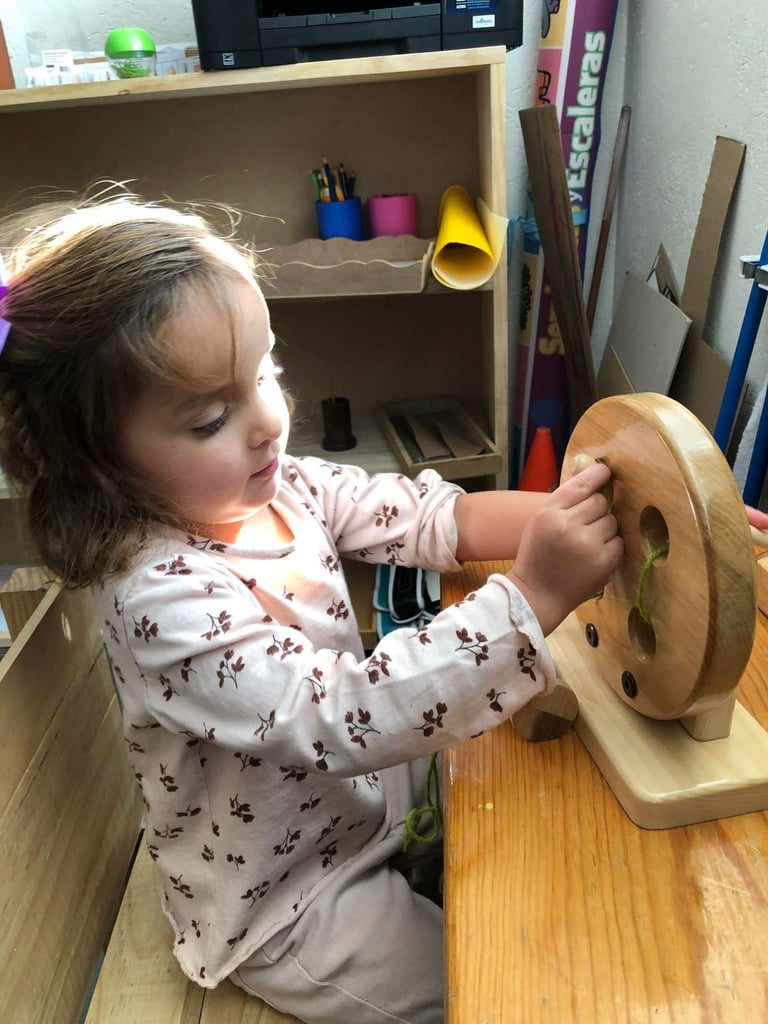Toddler in floral shirt playing with a wooden toy with holes at a classroom table near supply shelves