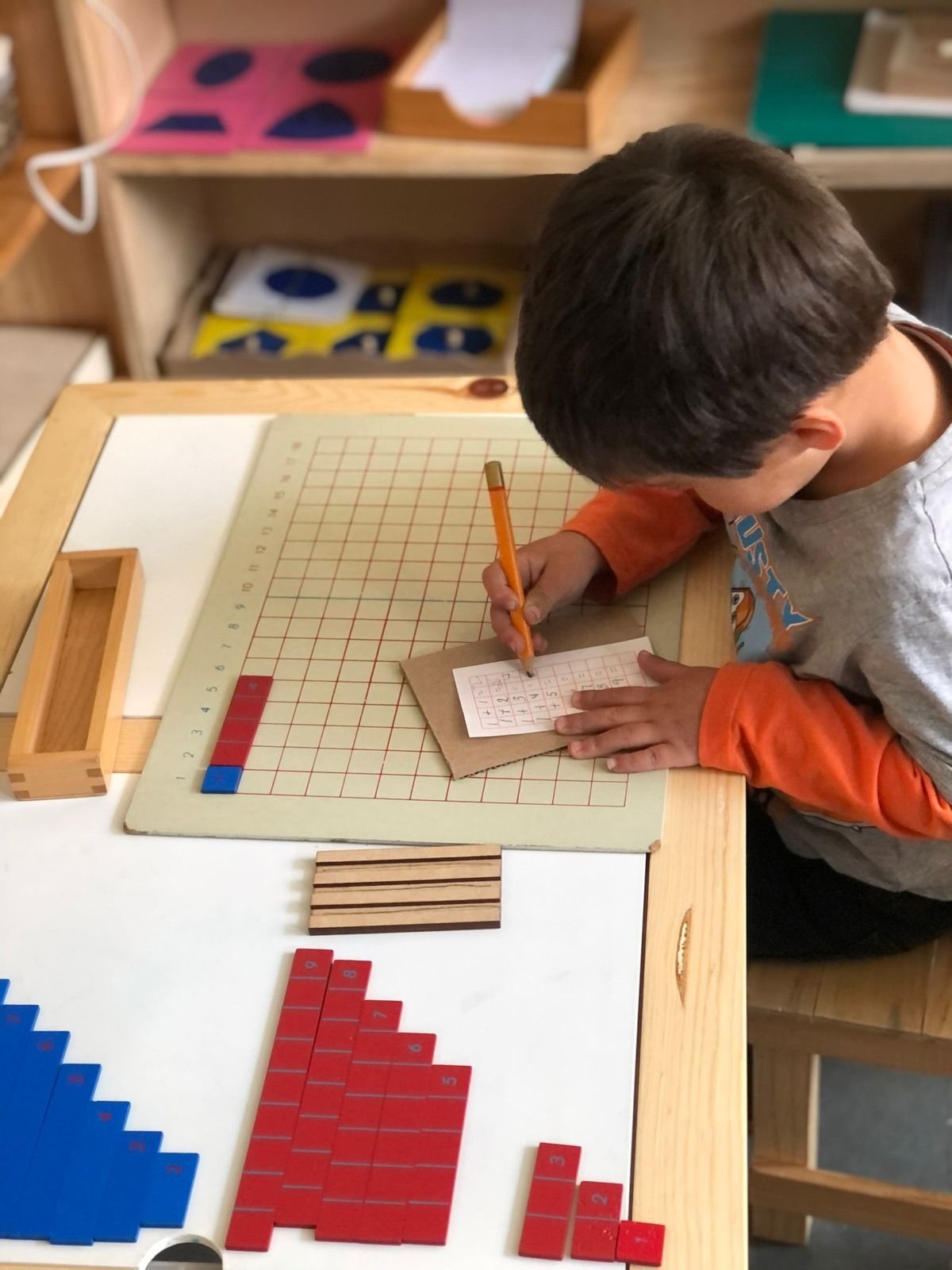 Child in orange jacket working on math with grid paper and wooden blocks at a learning table with educational materials