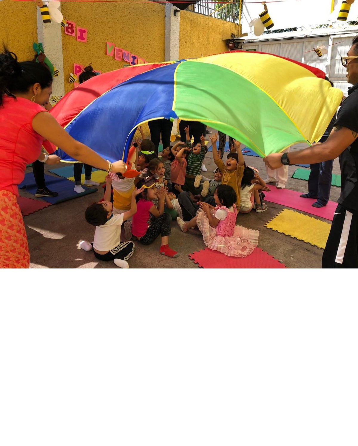 Children playing under a colorful rainbow parachute held by adults during an outdoor group activity at a school or community event.