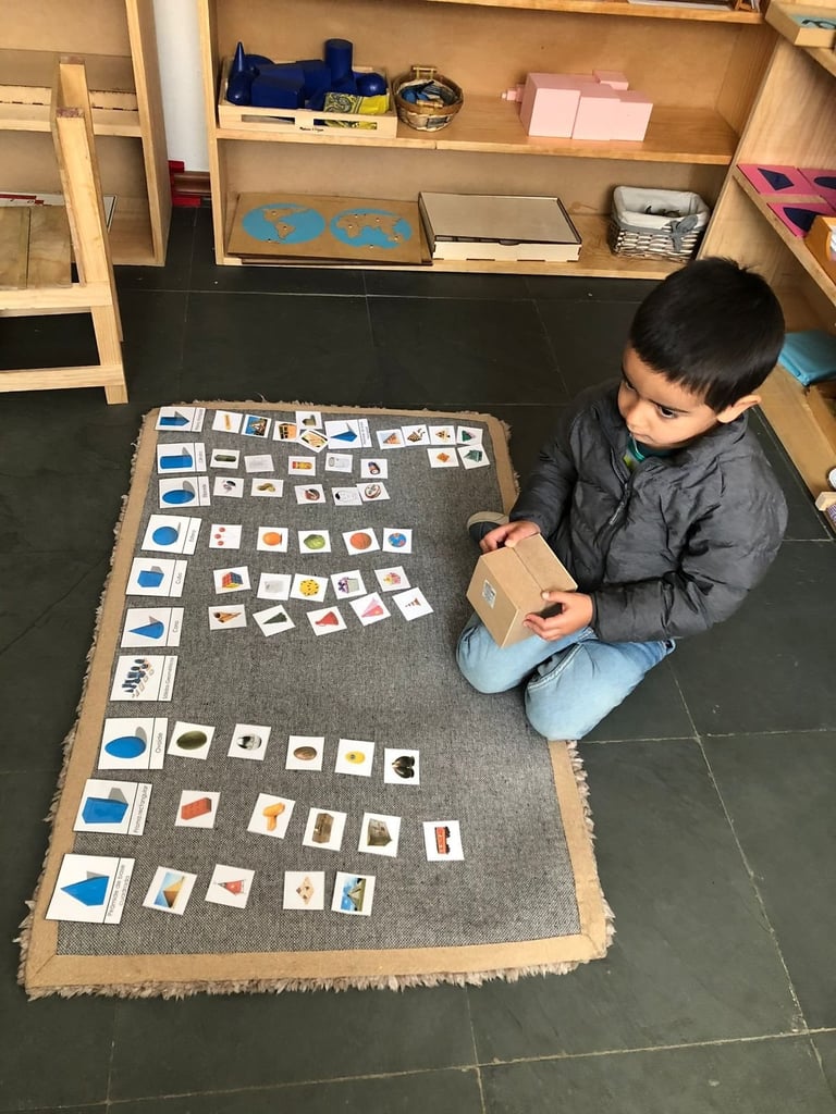 Child sitting on classroom floor engaged with educational cards and materials laid out on a mat, with wooden shelving visible in background.